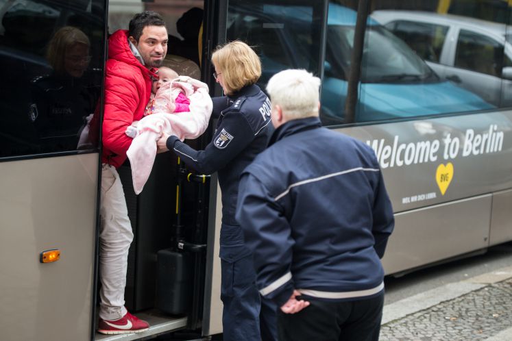 Eine Polizistin hilft einem Asylbewerber und seinem Baby am 11.03.2016 vor Registrierungszentrum des Berliner Landesamts für Gesundheit und Soziales (LaGeSo) in Berlin beim Einsteigen in einen Bus, auf dem "Welcome to Berlin" steht