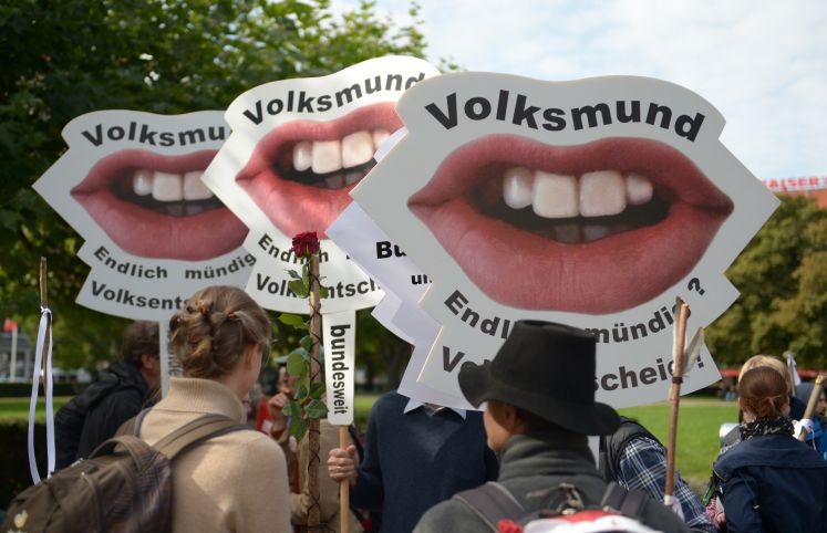 Demostranten mit Protestschildern, auf denen jeweils ein Mund zu sehen ist. Der Schriftzug auf den Plakten lautet: Volksmund. Endlich mündig? Volksentscheid!