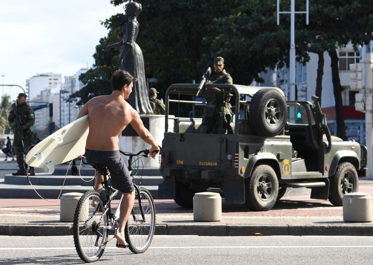 Ein Surfer fährt an der Copacabana von Rio de Janeiro an einem Jeep der Armee mit einem Soldaten, der ein Gewehr in der Hand hält, vorbei