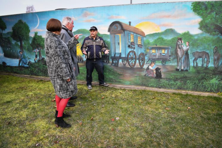 Drei Menschen besprechen ein jenisches Kulturzentrum. Hinter ihnen ist eine bemalte Wand mit eine historische Szene der Jenischen mit Pferden und Wagons