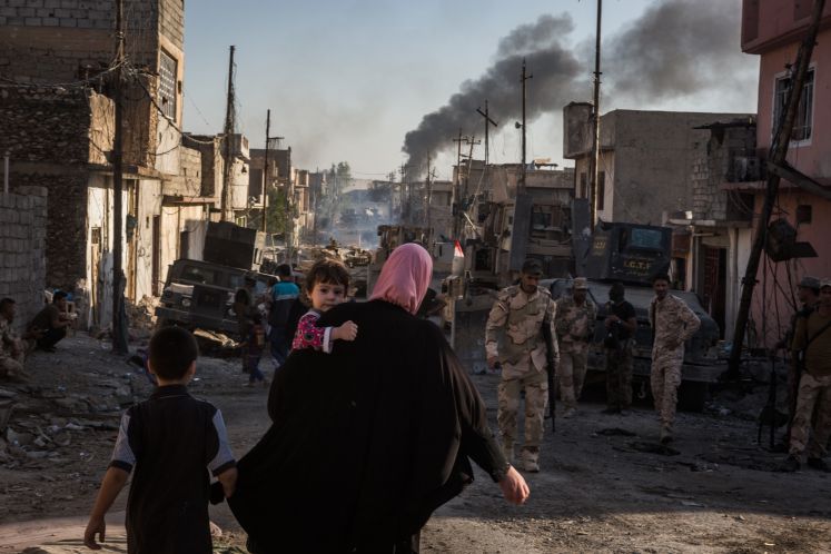 Eine Frau mit Kleinkind auf dem Arm und einem kleinem Jungen neben ihr, läuft in Mossul den irakischen Soldaten entgegen. Die Straßenzüge sind zerstört. Dunkler Rauch steigt am Ende der Straße auf