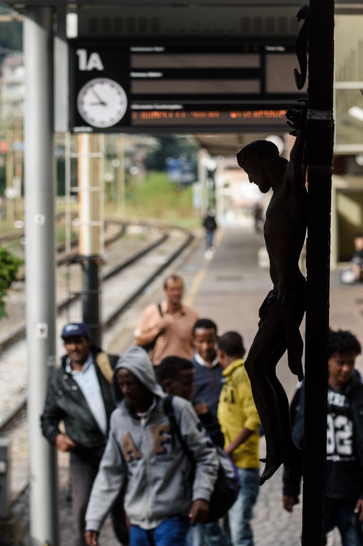 Zahlreiche Flüchtlinge gehen am 19.08.2015 auf einem Bahnsteig in Bozen (Italien) an einer am Kreuz hängenden Jesusfigur entlang 
