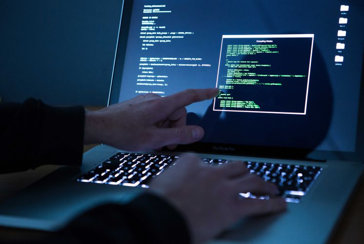 Ein Mann bedient eine beleuchtete Tastatur eines Laptops. Er zeigt auf den Monitor, auf dem ein Hacker-Programm geöffnet ist.