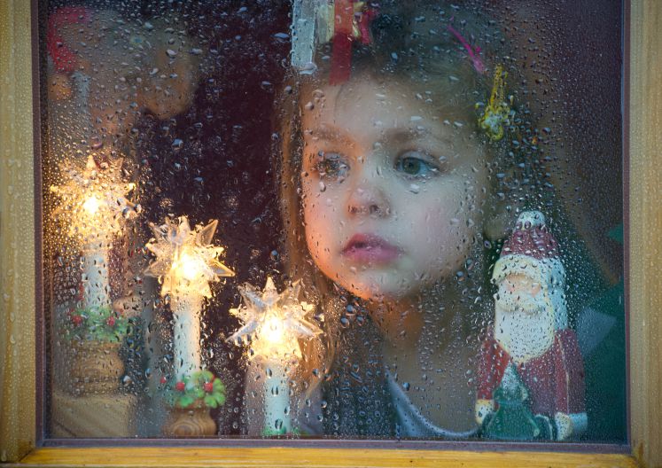Ein Kind schaut aus einem verregneten Fenster nach draußen, daneben Weihnachtsdekoration 