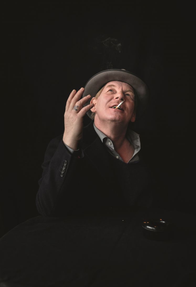 Ben Becker vor schwarzem Hintergrund mit Zigarette im Mund, die rechte Hand und seinen Blick nach oben gerichtet