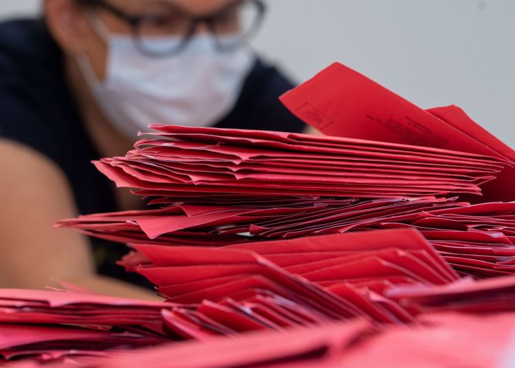 Stapel von roten Briefwahlumschlägen, im Hintergrund Mensch mit Maske