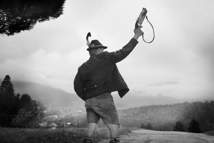 Ein Mann mit bayerischer Lederhose, Hut und Jacke schießt mit einem Handböller vor einem Bergpanorama in die Luft