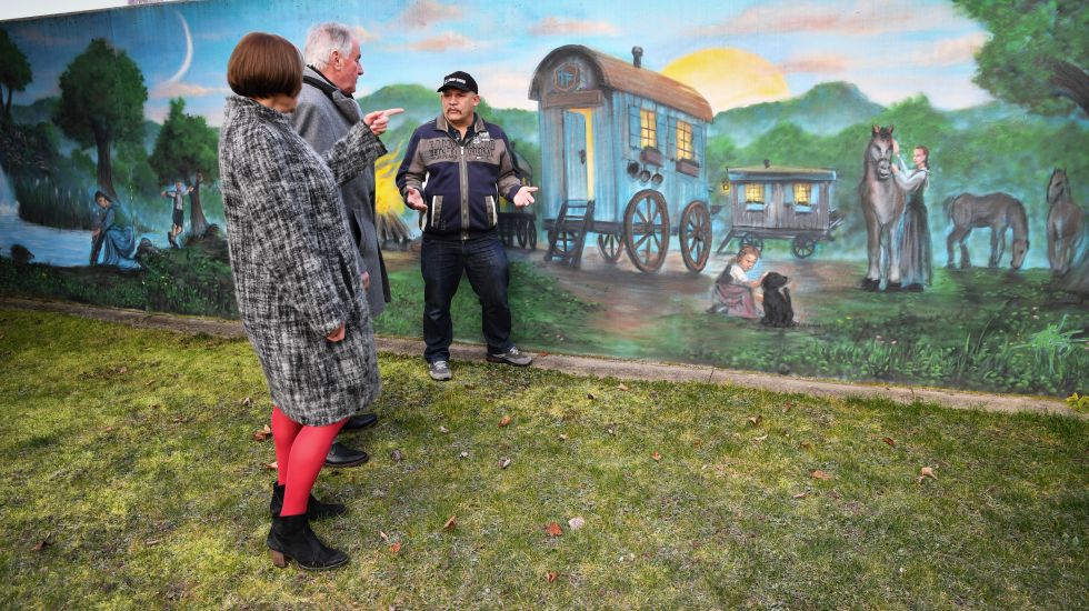 Drei Menschen besprechen ein jenisches Kulturzentrum. Hinter ihnen ist eine bemalte Wand mit eine historische Szene der Jenischen mit Pferden und Wagons