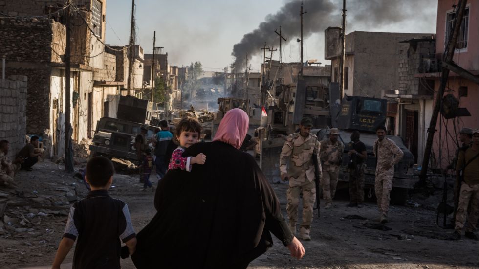 Eine Frau mit Kleinkind auf dem Arm und einem kleinem Jungen neben ihr, läuft in Mossul den irakischen Soldaten entgegen. Die Straßenzüge sind zerstört. Dunkler Rauch steigt am Ende der Straße auf