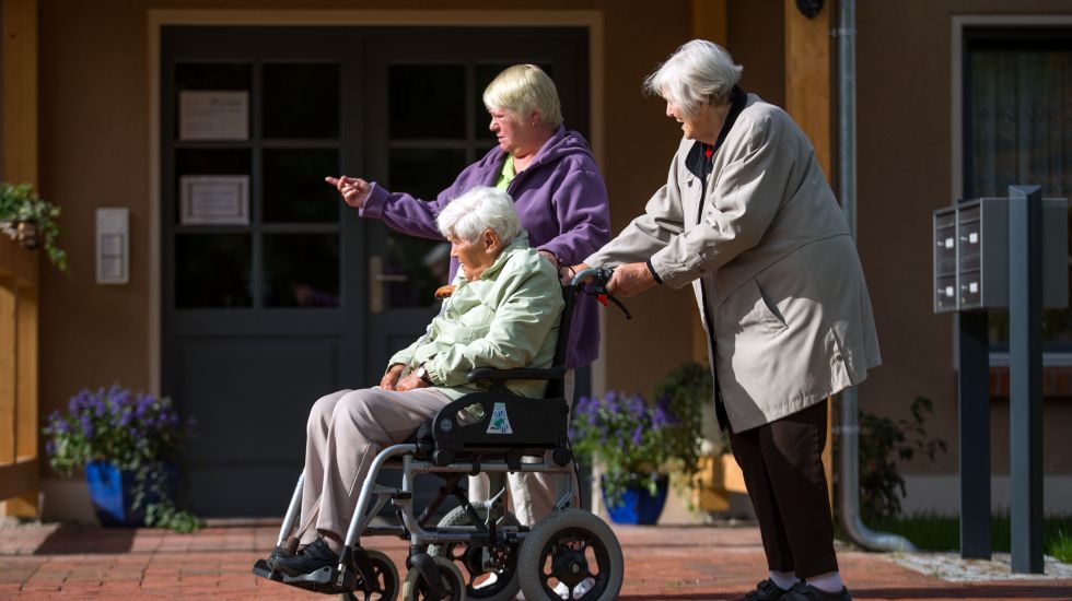 Zwei ältere Damen schieben eine alte Dame im Rollstuhl