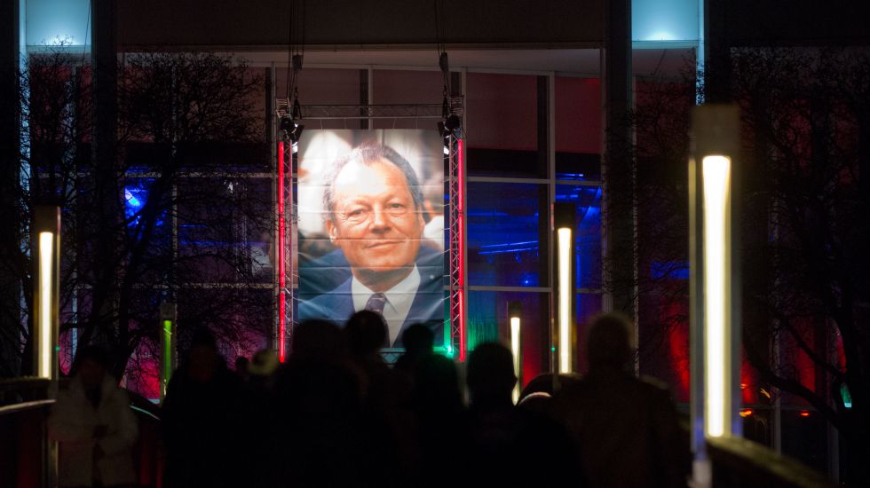 Passanten gehen am 11.12.2013 über eine Brücke in Richtung der Musik- und Kongresshalle in Lübeck (Schleswig-Holstein) an der ein Bild von Willy Brandt hängt.