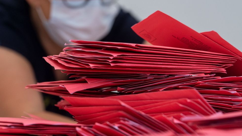 Stapel von roten Briefwahlumschlägen, im Hintergrund Mensch mit Maske