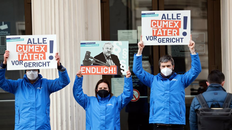 Aktivisten der „Bürgerbewegung Finanzwende jetzt“ halten vor dem Landgericht Plakate mit der Aufschrift „Alle Cumex Täter vor Gericht“